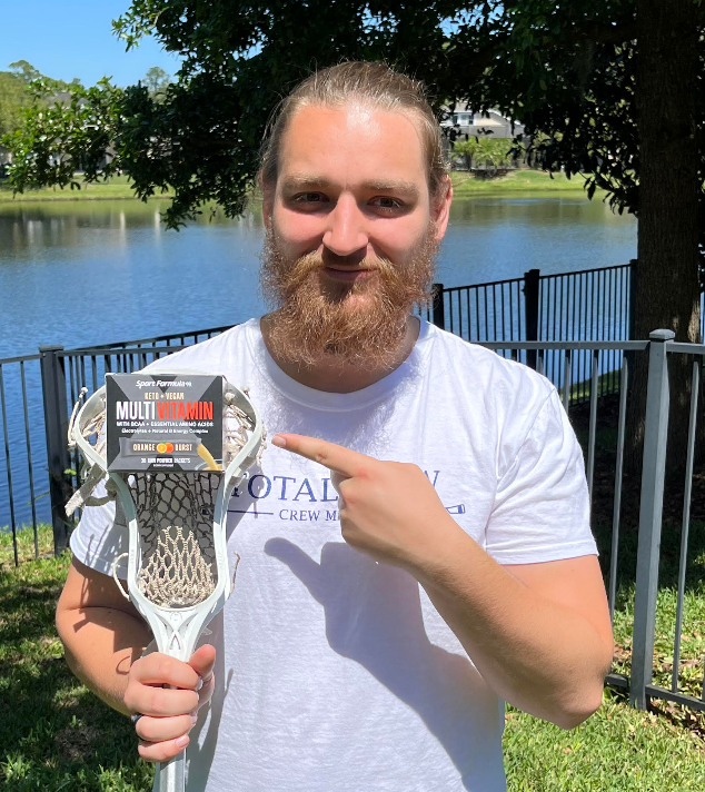 A person holds a lacrosse stick with a multivitamin package while standing by a serene lake surrounded by greenery.