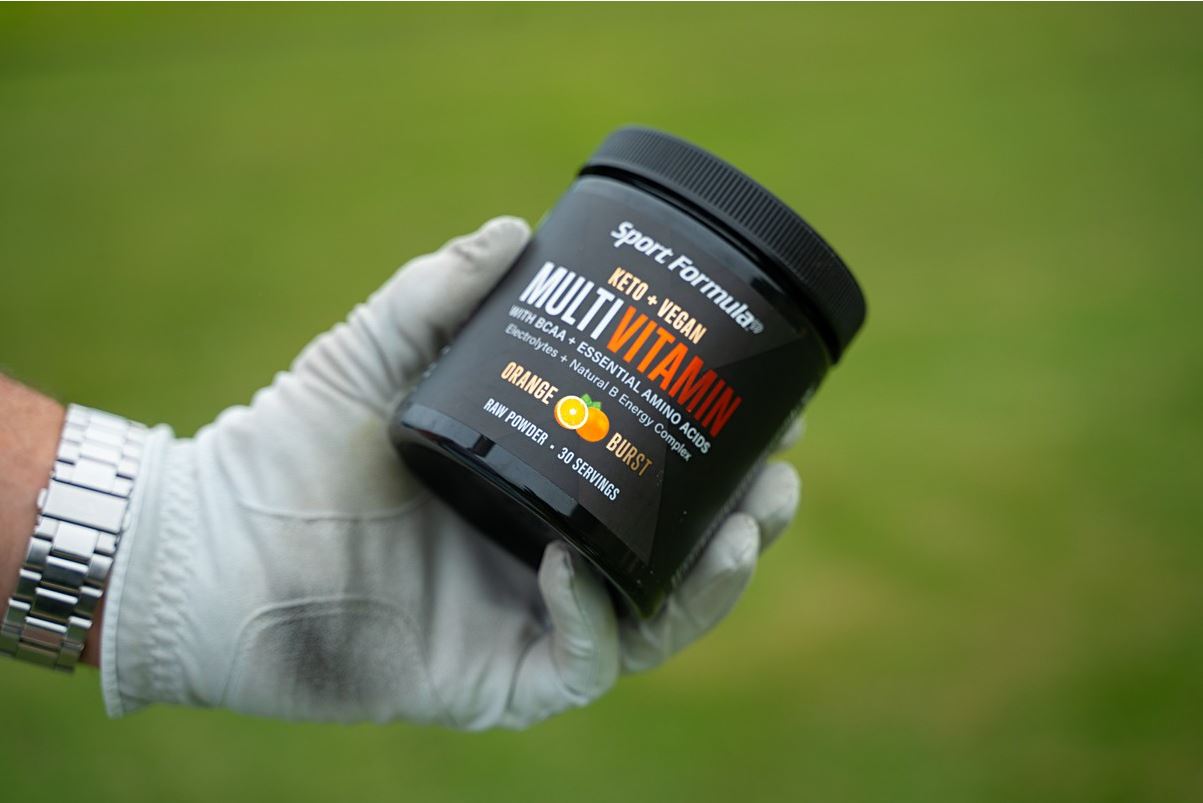 A gloved hand holds a black jar of Sport Formula Multivitamin, labeled Keto + Vegan in orange, with an orange burst flavor.
