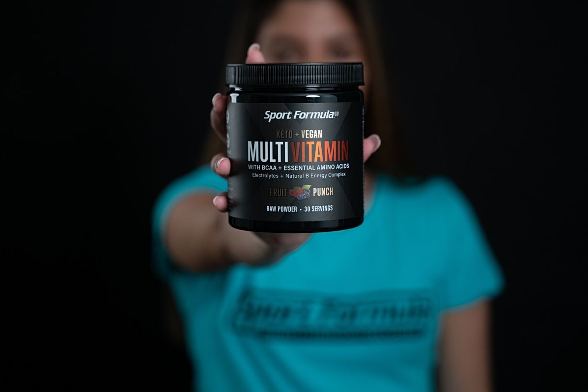 A hand holds a container of Multi Vitamin with BCAA, labeled Fruit Punch, against a black background. The person is wearing a blue shirt.