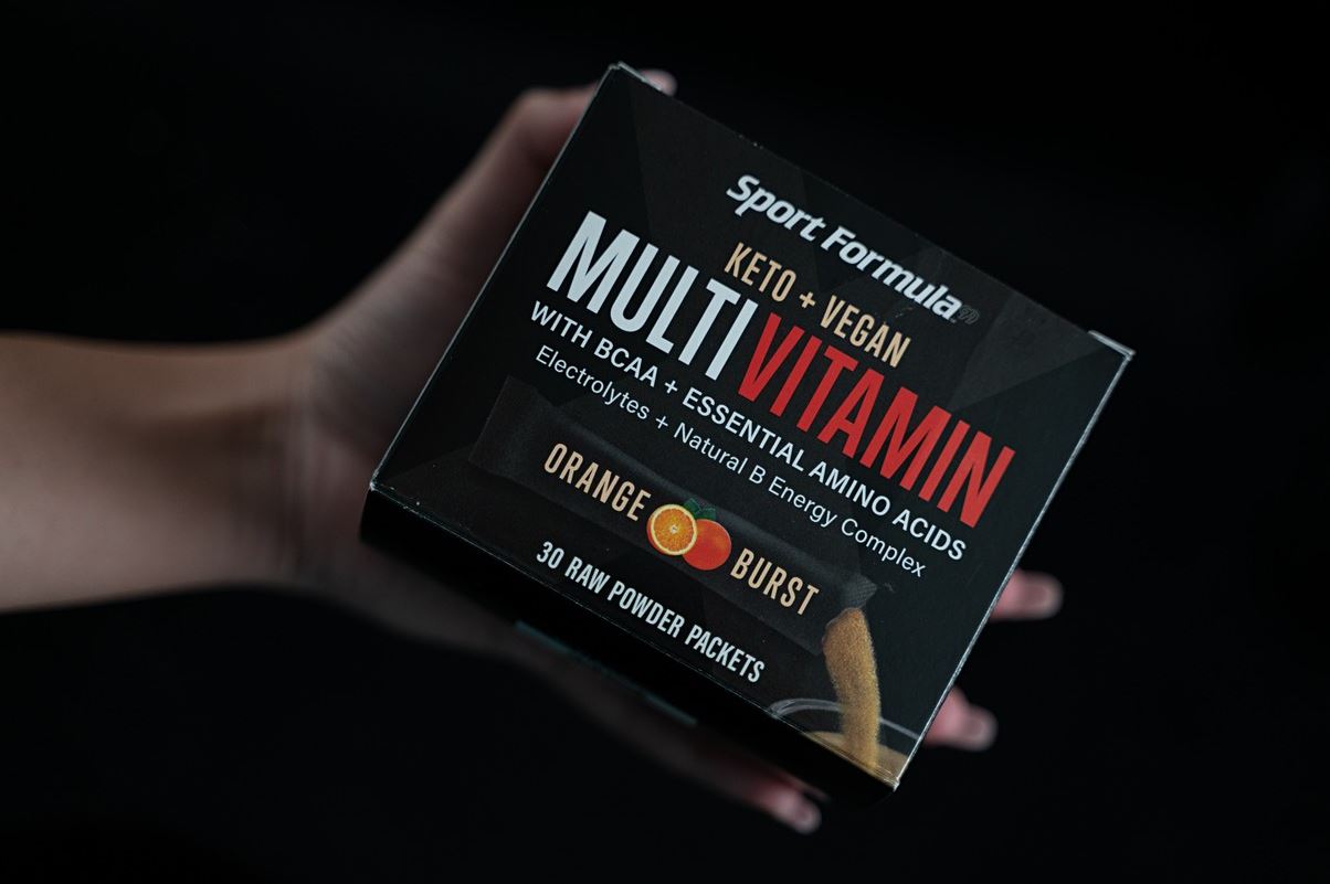 A hand holds a box of Sport Formula Keto + Vegan Multivitamin with BCAA and electrolytes, featuring 30 orange burst powder packets.