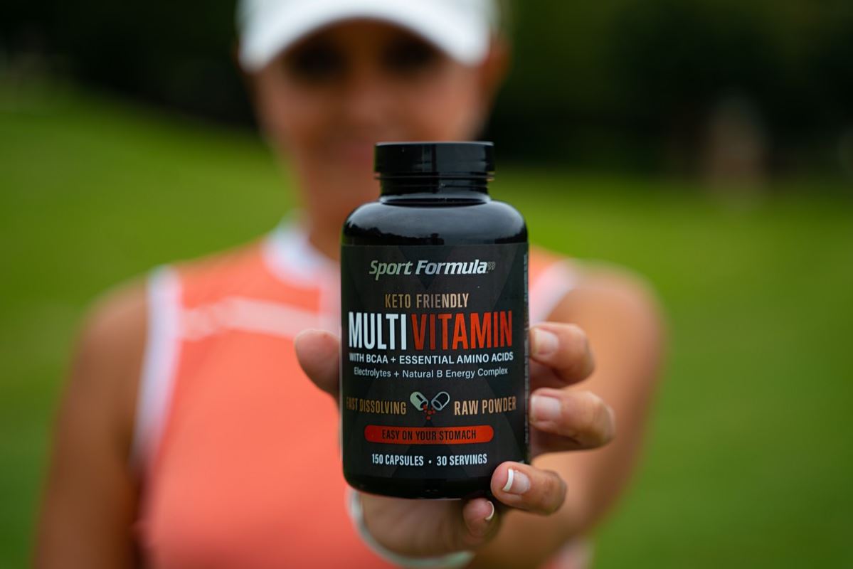 A person holds a black container of Sport Formula Keto Friendly Multivitamin, promoting BCAAs and essential amino acids outdoors.