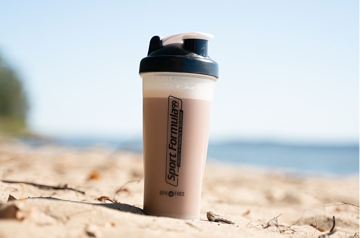 A protein shaker sits on sandy beach with a blurred ocean background, labeled Sport Formula 99 for energy and recovery.