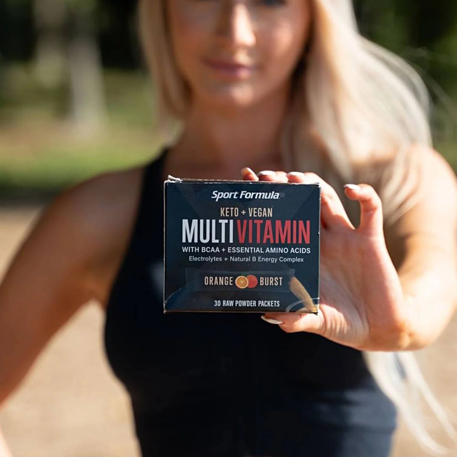 A person holds a box of Sport Formula MultiVitamin, labeled keto and vegan, featuring orange burst flavor and essential amino acids.