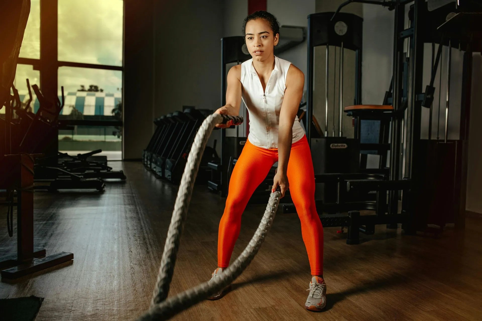 A fit individual is performing an exercise with battle ropes in a gym, wearing a white top and bright orange leggings.