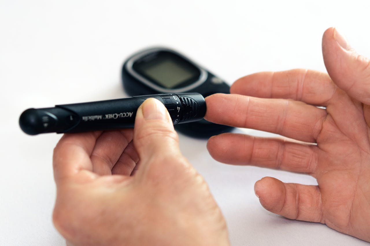 A person uses a blood glucose lancing device on their fingertip, with a glucose meter visible in the background.