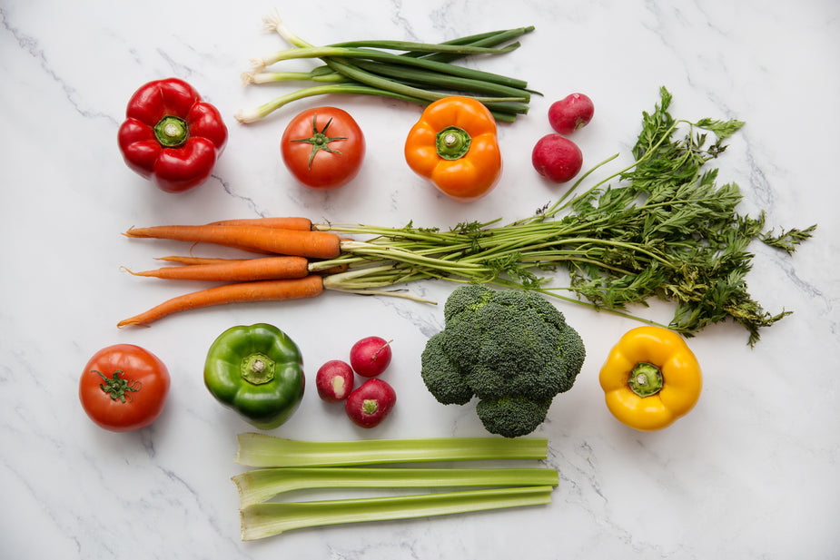 A nutrient colorful assortment of fresh vegetables including carrots, tomatoes, bell peppers, broccoli, radishes, celery, and spring onions on marble.