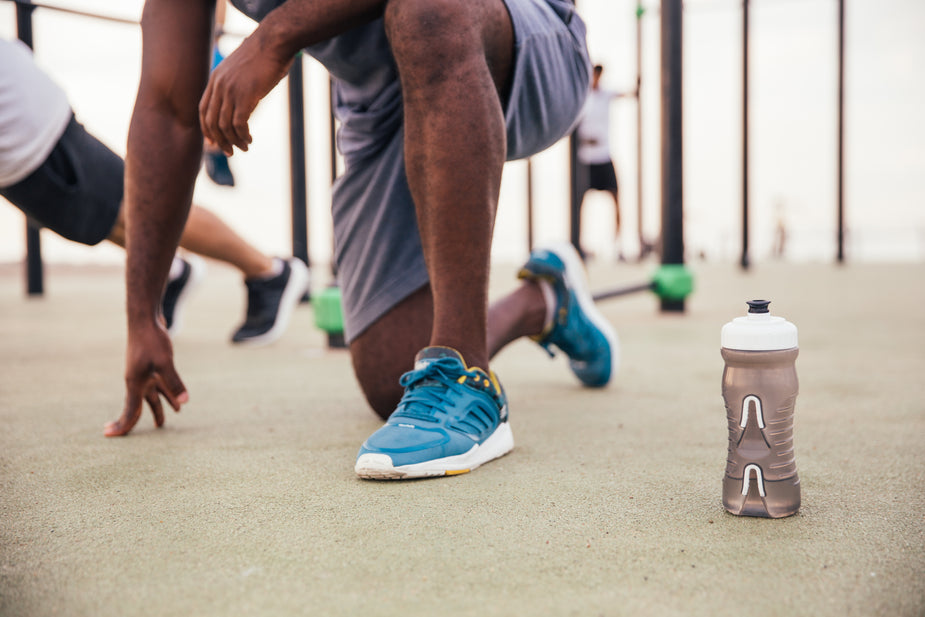 A close-up of a crouching athlete in blue sneakers, preparing to exercise, with a water bottle beside him in an outdoor gym setting.