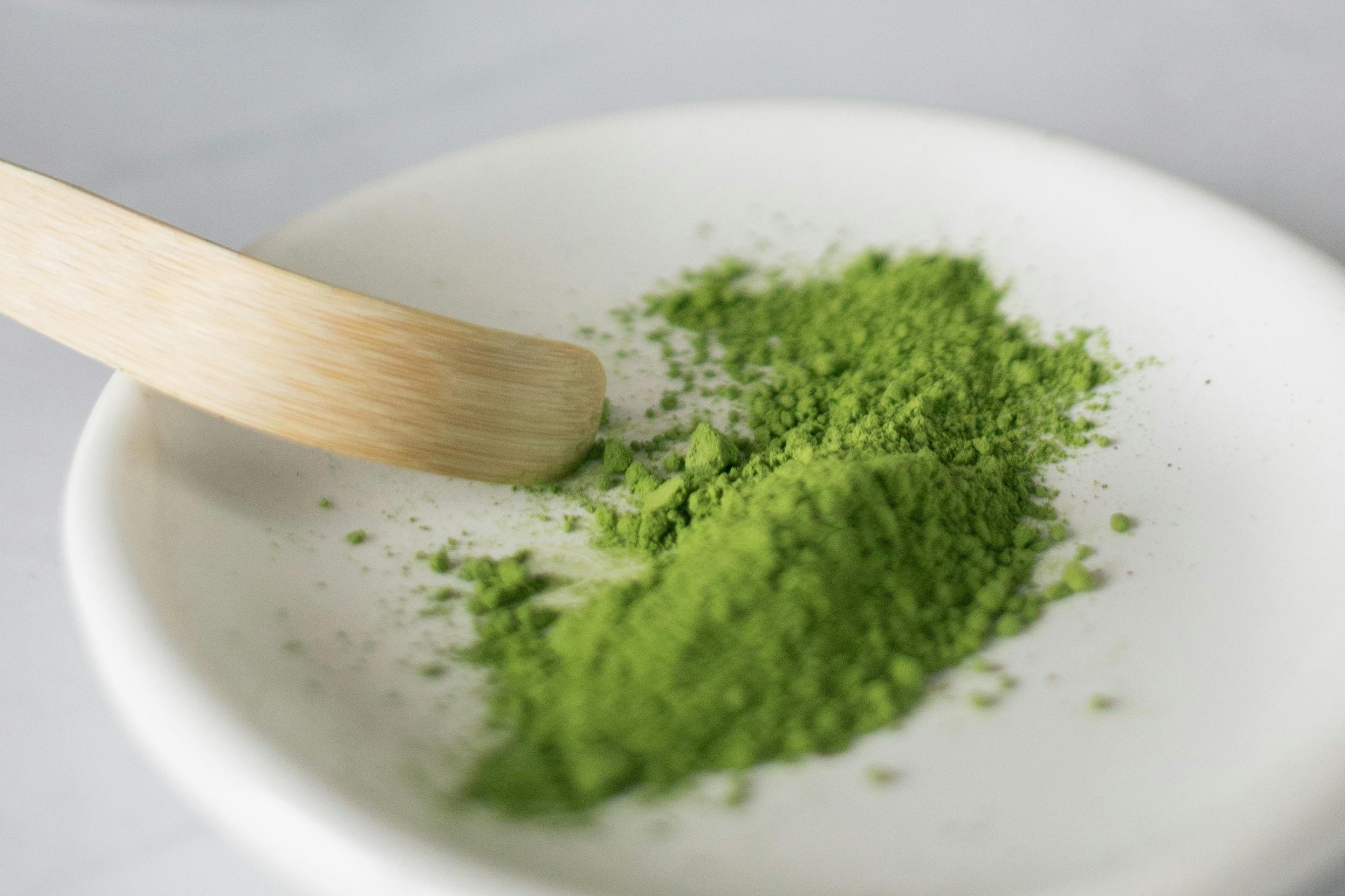 A small white dish holds vibrant green powder, with a wooden scoop nearby, ready for use in tea or recipes.