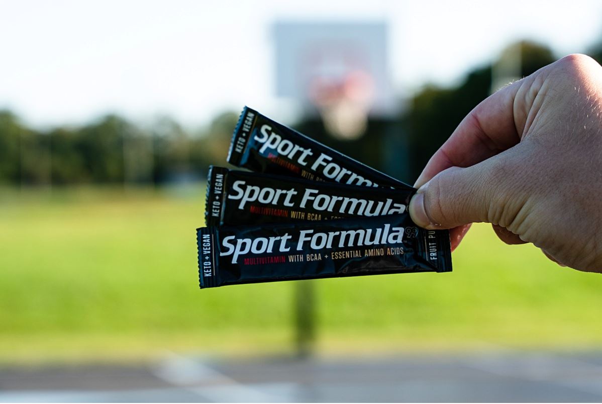 A hand holds three packets of Sport Formula vitamin supplements with a basketball hoop in the background on a sunny day.