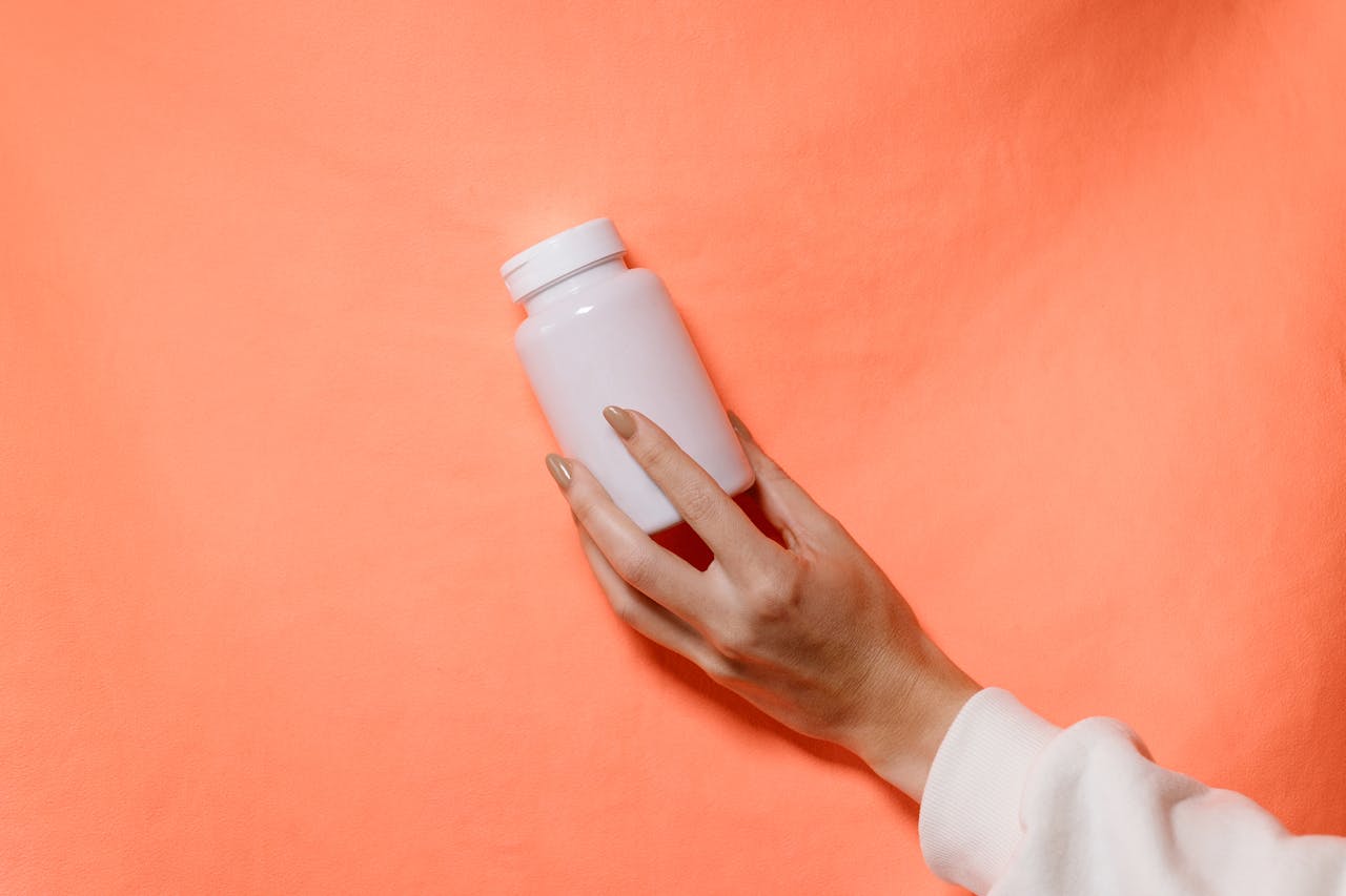 A hand holds a white supplement bottle against a bright coral background. The focus is on the bottle and the hand's elegant grip.