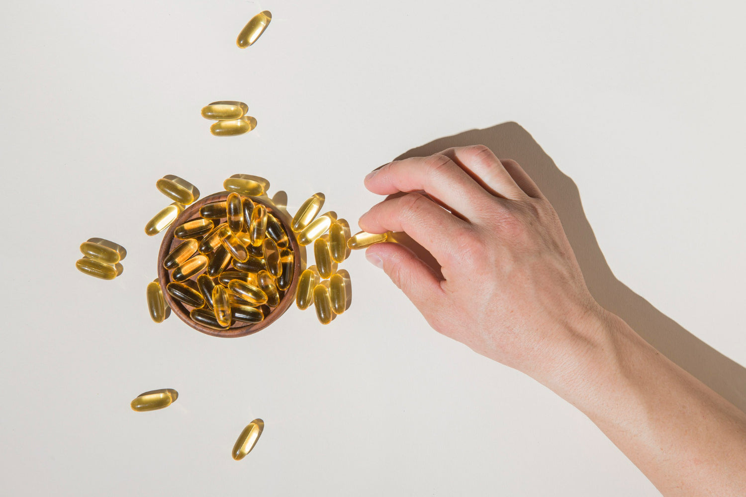 A hand reaching for a golden gel capsule from a wooden bowl surrounded by scattered capsules on a bright surface.