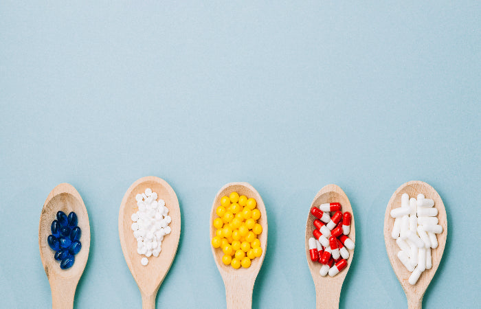 Five wooden spoons holding different colored pills and capsules against a light blue background.