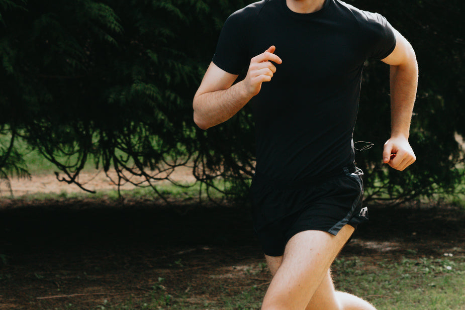 A man in a black athletic shirt and shorts runs through a park, surrounded by lush green trees.