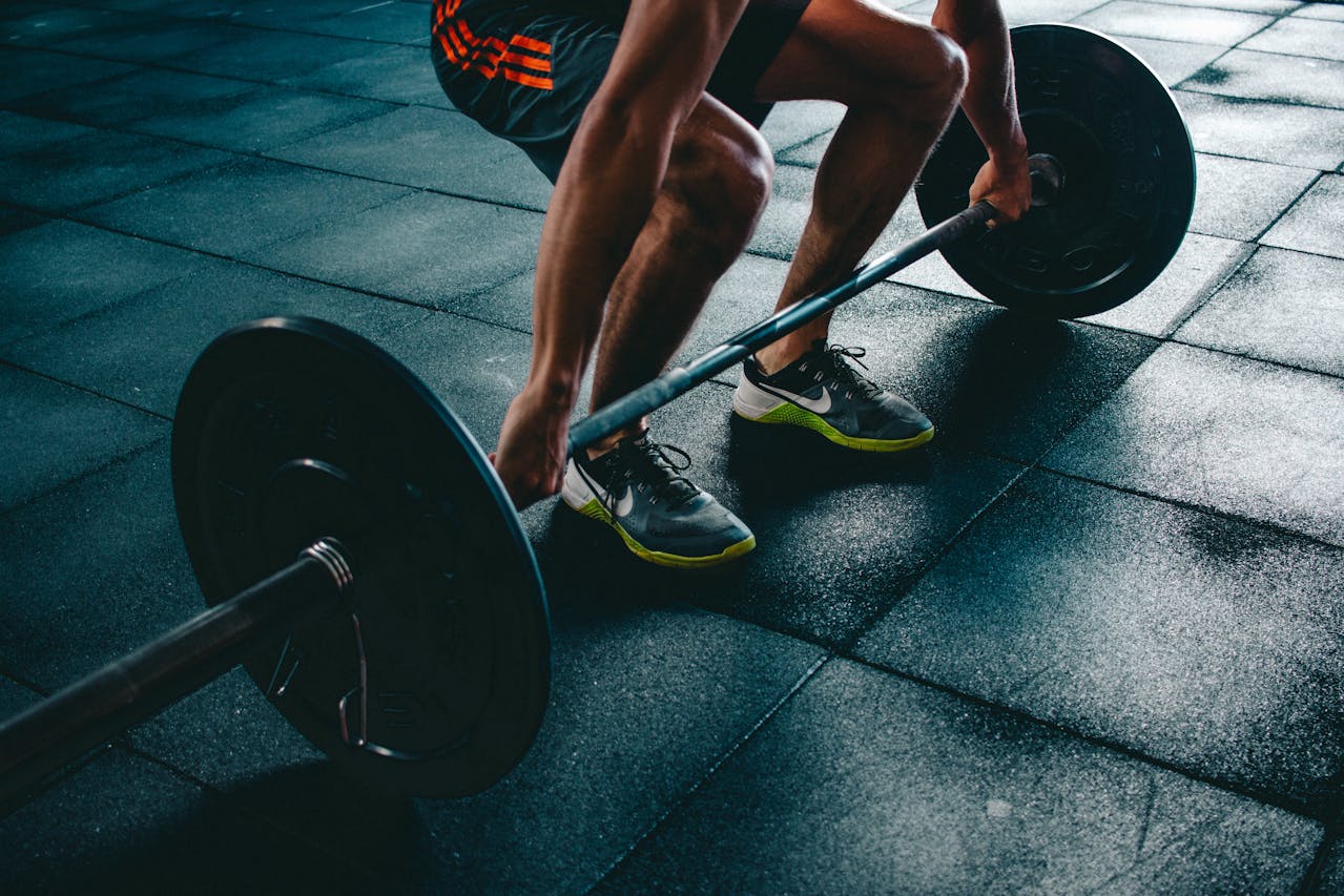 A close-up of a person lifting a barbell with weights, showcasing muscular legs and athletic shoes on a gym floor.