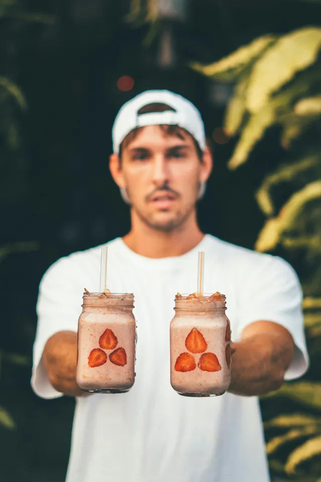 A person holds two jars filled with creamy strawberry smoothies, each with decorative fruit slices, against a lush green backdrop.