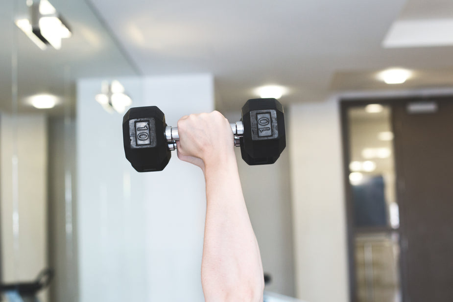 A raised hand holding a 10-pound dumbbell in a gym setting, with mirrors and soft lighting in the background.