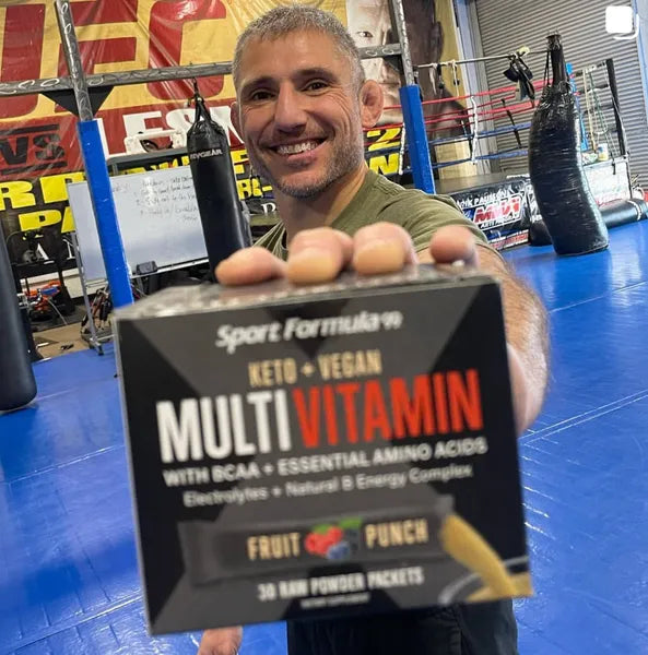 A person holds a box of Sport Formula multi-vitamins labeled Keto Vegan and Fruit Punch in a gym with boxing bags in the background.