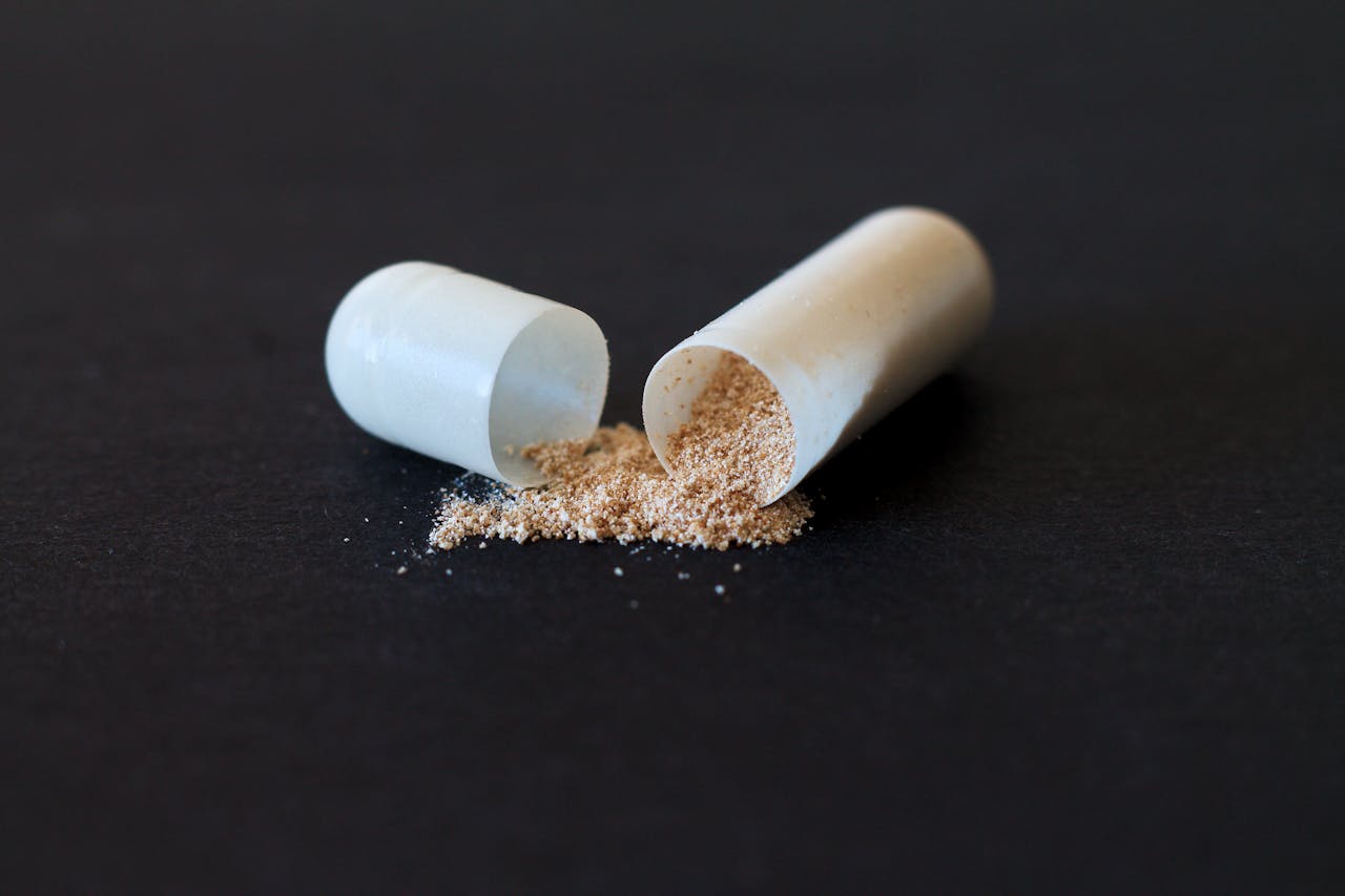 A split open capsule lies on a dark surface, spilling out fine, light brown powder.