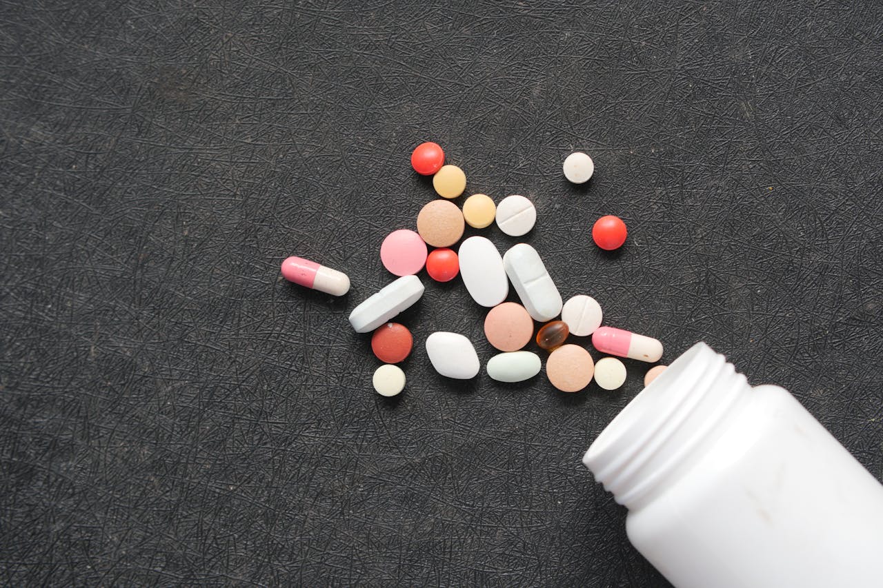 A supplement bottle spills out various colored pills and capsules onto a dark, textured surface.