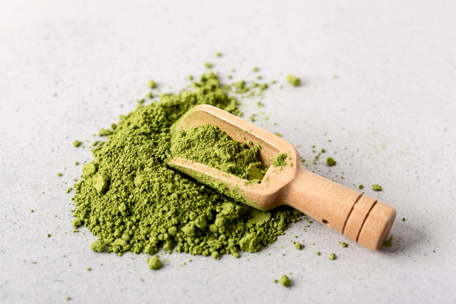 Green powder with a wooden scoop resting on top, set against a light gray background. Ideal for wellness or culinary use.