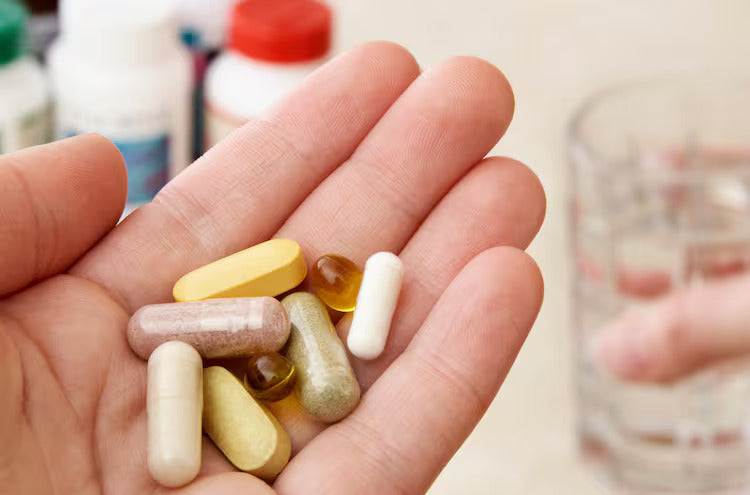 A hand holding assorted supplements with a blurry glass of water in the background.