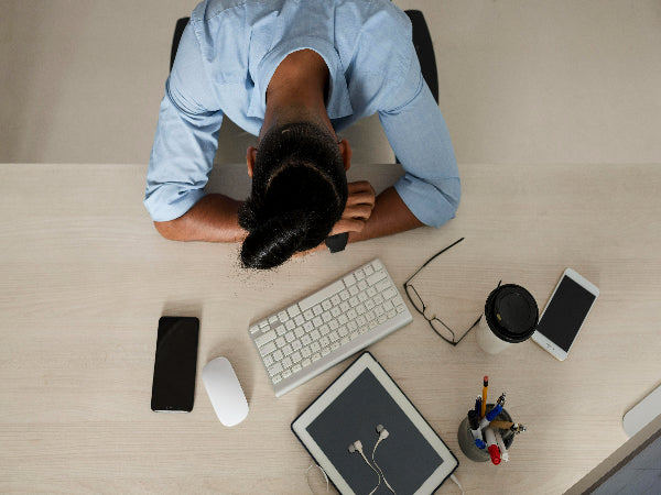 Person with head on desk, surrounded by a keyboard, smartphone, tablet, mouse, coffee cup, and glasses.