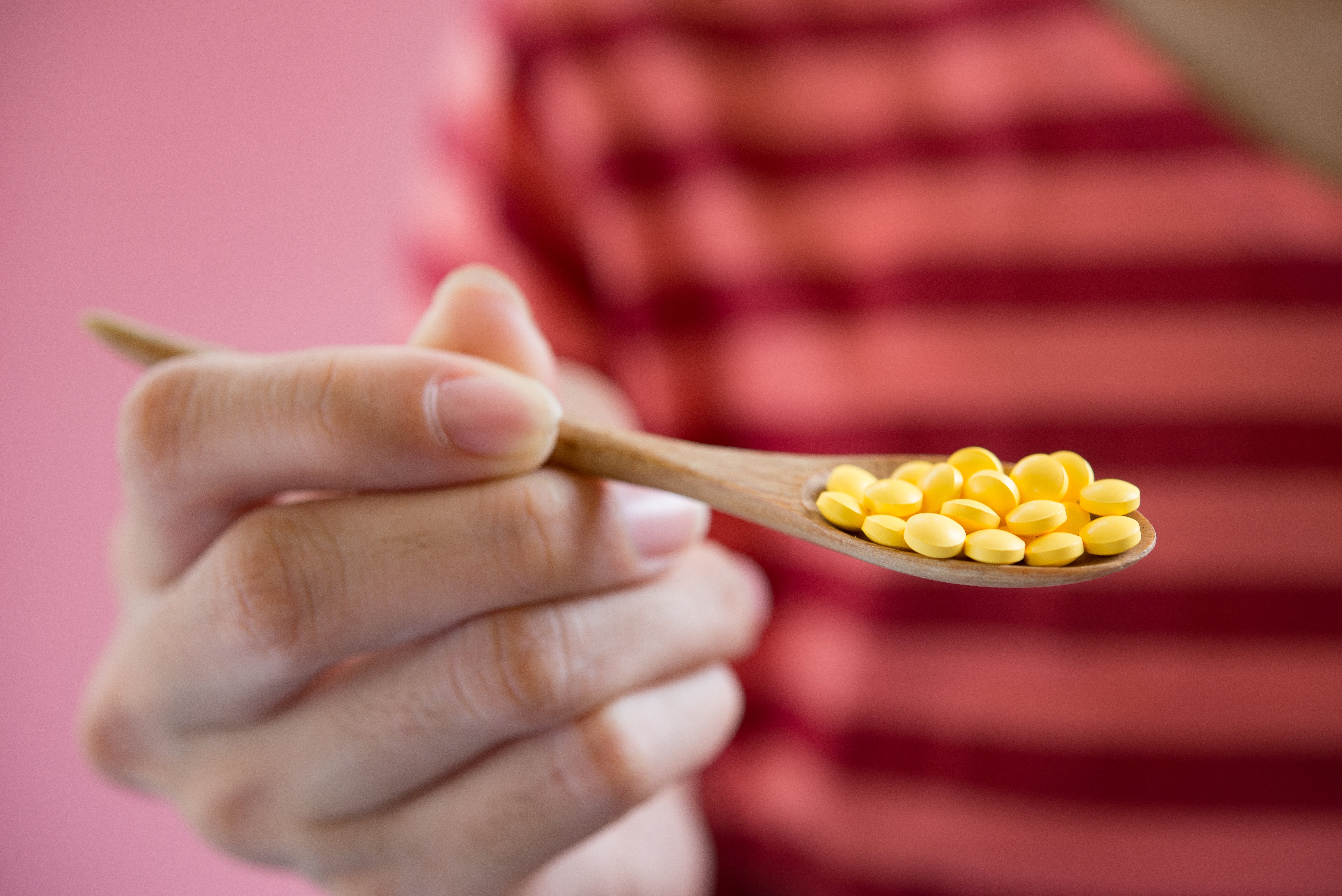 Person holding a wooden spoon full of yellow pills with a pink background.