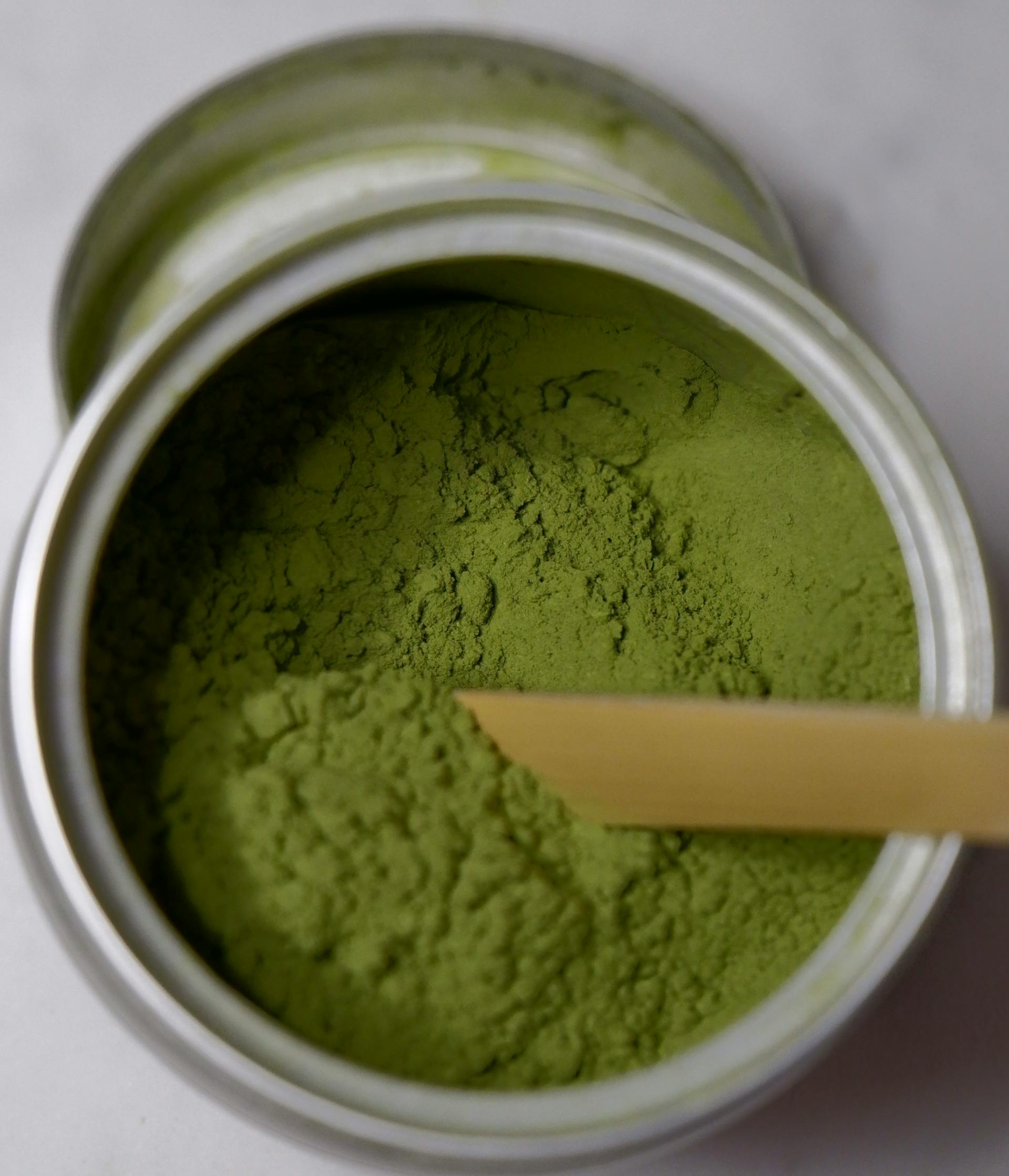 A close-up view of a tin containing vibrant green powder, with a bamboo scoop partially submerged in the matcha-like substance.