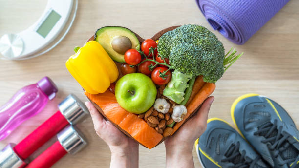 Hands holding a heart-shaped board with assorted healthy foods, surrounded by fitness gear.