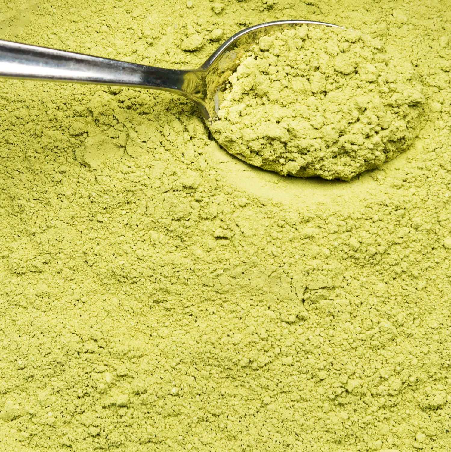 A close-up of vibrant green powdered substance, with a silver spoon scooping a portion from a larger pile of the fine powder.
