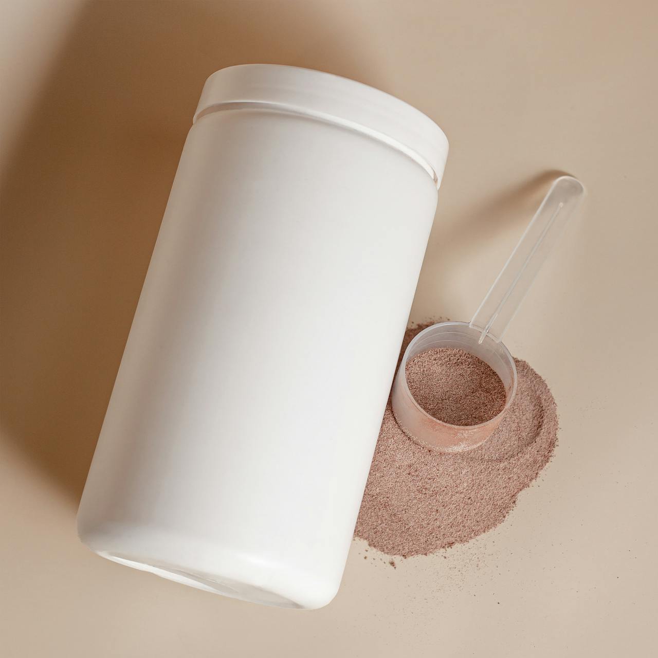 A white cylindrical container lies on its side, next to a mound of brown powder and a transparent scoop on a beige background.