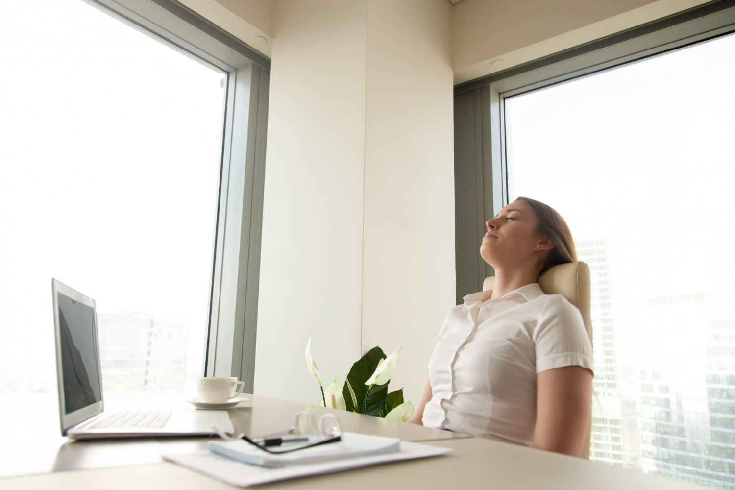 A woman in a white blouse rests in an office chair, overlooking a city skyline through large windows with a laptop open on her desk.