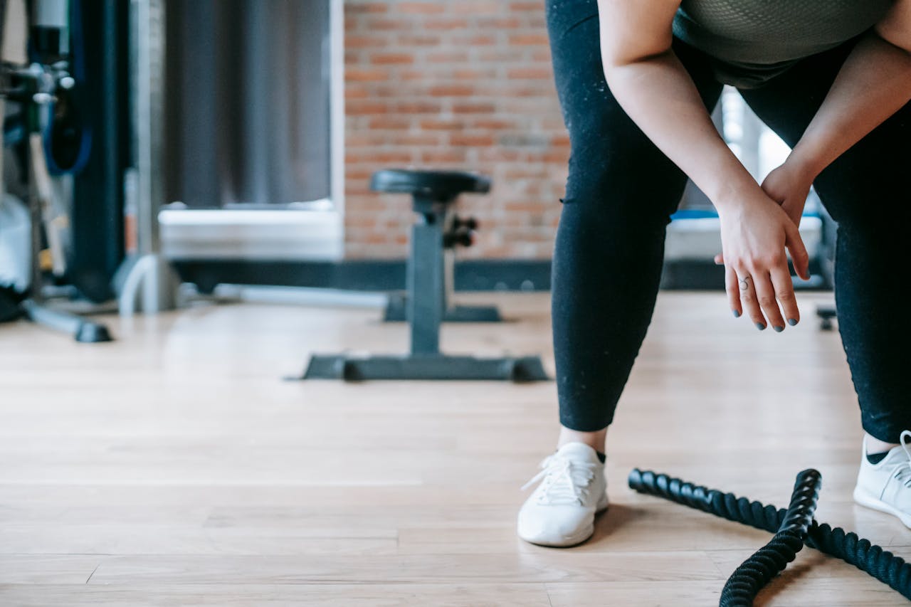 A person in workout clothes takes a break, hands on knees, beside battle ropes on a gym floor with a brick wall in the background.